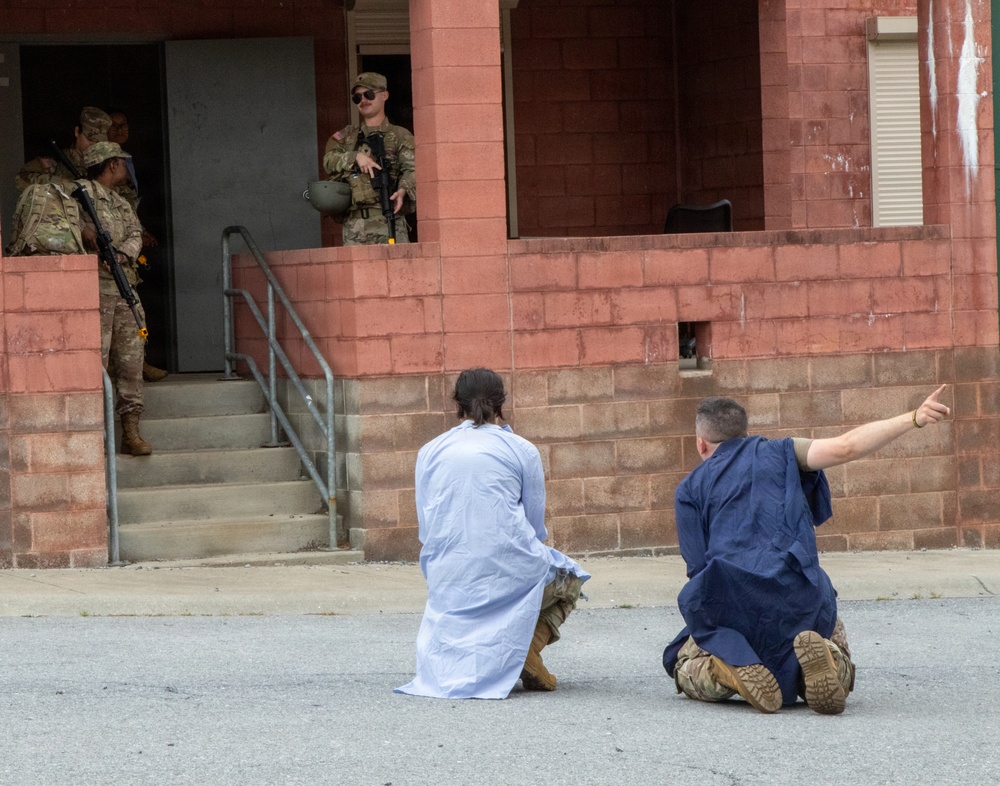The image depicts what appears to be a military training exercise involving role-players in civilian-style robes kneeling on the ground in front of armed soldiers in full combat gear. Two individuals in loose garments are positioned on their knees facing a building entrance where armed personnel in camouflage uniforms are standing. The setting is a red brick/cinder block building with steps, consistent with a military installation training facility.