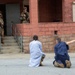 The image depicts what appears to be a military training exercise involving role-players in civilian-style robes kneeling on the ground in front of armed soldiers in full combat gear. Two individuals in loose garments are positioned on their knees facing a building entrance where armed personnel in camouflage uniforms are standing. The setting is a red brick/cinder block building with steps, consistent with a military installation training facility.