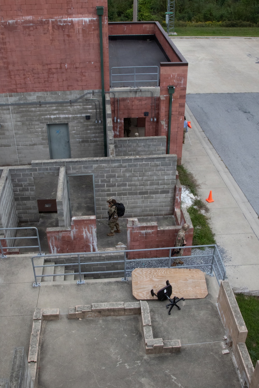 An aerial/overhead view of what appears to be a military or law enforcement training facility with mock building structures made of concrete block walls. Several personnel in tactical gear are navigating through the roofless training rooms, while a person in civilian clothing sits in an office chair near a plywood board at the lower level. Orange traffic cones and metal railings are visible around the perimeter.