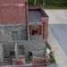 An aerial/overhead view of what appears to be a military or law enforcement training facility with mock building structures made of concrete block walls. Several personnel in tactical gear are navigating through the roofless training rooms, while a person in civilian clothing sits in an office chair near a plywood board at the lower level. Orange traffic cones and metal railings are visible around the perimeter.