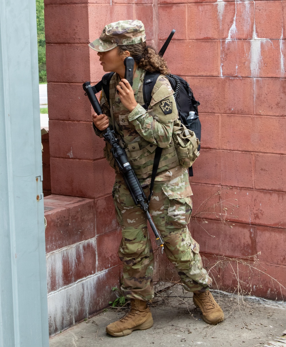 A female soldier in U.S. Army OCP (Operational Camouflage Pattern) uniform is positioned against a red brick wall, appearing to conduct a radio communication while holding a rifle. She is equipped with a tactical backpack, a radio handset with a coiled cord, and carries what appears to be an M4/M16-style rifle. The setting appears to be a military training environment or urban operations exercise.
