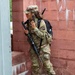 A female soldier in U.S. Army OCP (Operational Camouflage Pattern) uniform is positioned against a red brick wall, appearing to conduct a radio communication while holding a rifle. She is equipped with a tactical backpack, a radio handset with a coiled cord, and carries what appears to be an M4/M16-style rifle. The setting appears to be a military training environment or urban operations exercise.