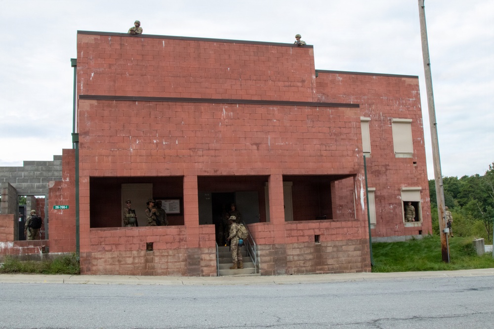 The image shows military personnel conducting what appears to be a training exercise at a mock urban environment facility. Soldiers in combat gear are positioned at multiple levels of a two-story red/pink cinder block building, including on the rooftop and at ground-floor entry points. The setting is consistent with a Military Operations in Urban Terrain (MOUT) training site.