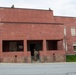 The image shows military personnel conducting what appears to be a training exercise at a mock urban environment facility. Soldiers in combat gear are positioned at multiple levels of a two-story red/pink cinder block building, including on the rooftop and at ground-floor entry points. The setting is consistent with a Military Operations in Urban Terrain (MOUT) training site.