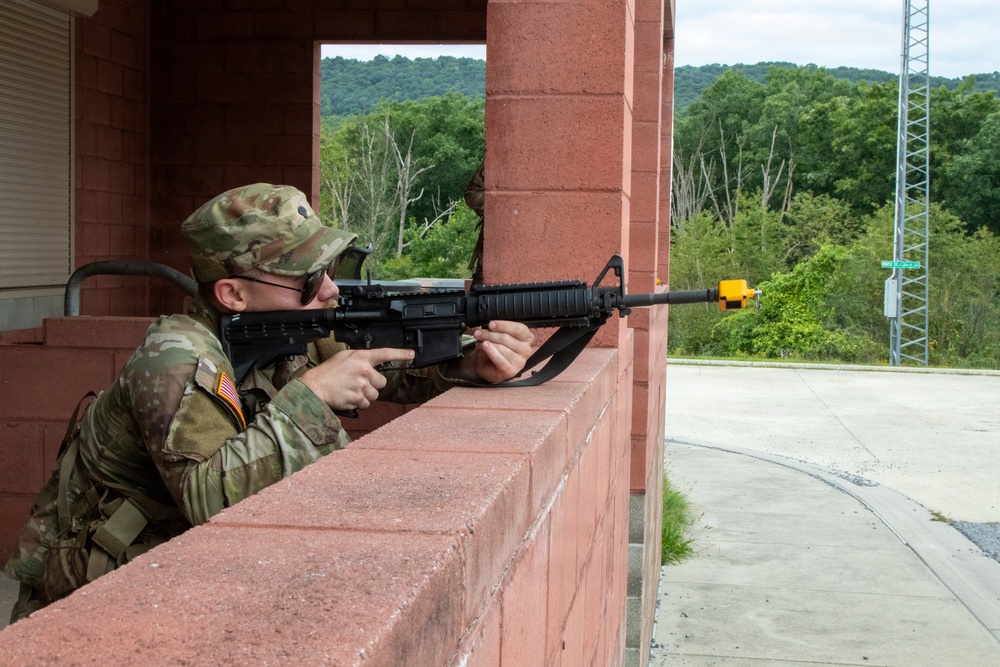 A U.S. Army soldier in MultiCam (OCP) camouflage uniform is positioned behind a red brick wall, aiming an M16/M4-style rifle with a yellow MILES (Multiple Integrated Laser Engagement System) device attached to the muzzle. The setting appears to be a military training facility, indicated by the MILES training device and the structured building environment. The soldier is practicing tactical shooting positions during what appears to be a field training exercise.