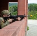 A U.S. Army soldier in MultiCam (OCP) camouflage uniform is positioned behind a red brick wall, aiming an M16/M4-style rifle with a yellow MILES (Multiple Integrated Laser Engagement System) device attached to the muzzle. The setting appears to be a military training facility, indicated by the MILES training device and the structured building environment. The soldier is practicing tactical shooting positions during what appears to be a field training exercise.