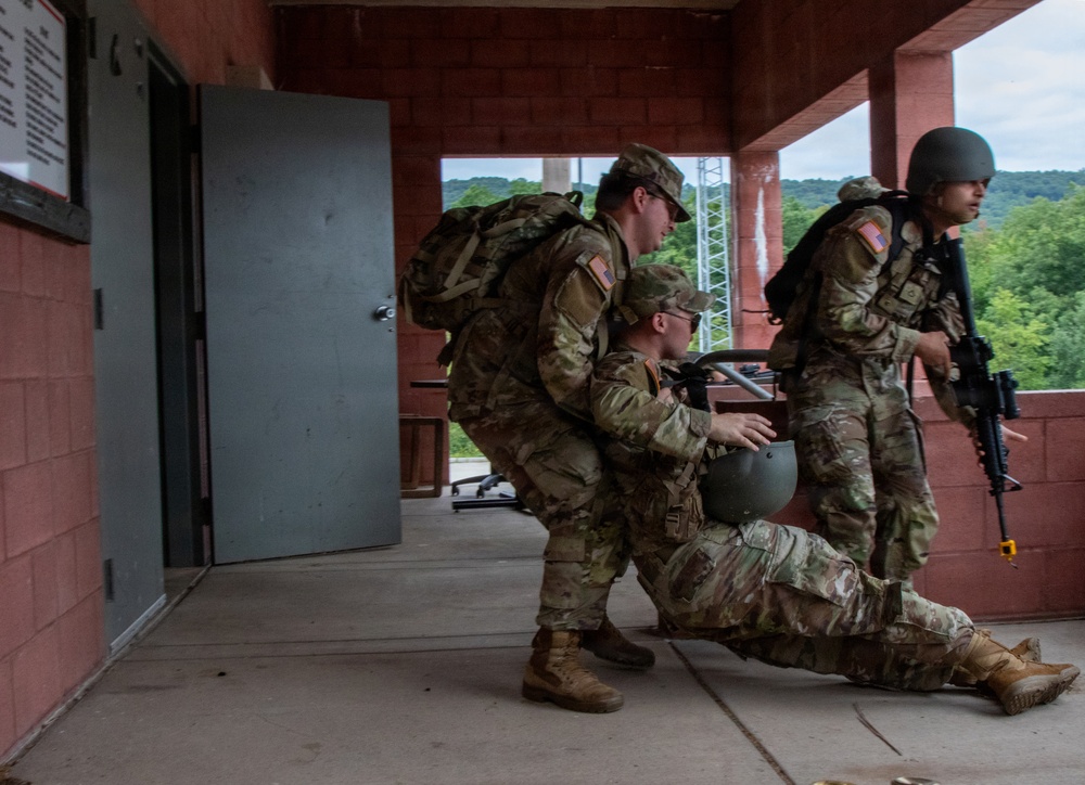 Three military personnel in MultiCam/OCP camouflage uniforms are conducting what appears to be a casualty evacuation (CASEVAC) drill on a covered concrete walkway of a red brick training facility. One soldier is being dragged or assisted along the ground while two others support and provide security. The setting appears to be a military training installation with wooded terrain visible in the background.
