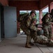 Three military personnel in MultiCam/OCP camouflage uniforms are conducting what appears to be a casualty evacuation (CASEVAC) drill on a covered concrete walkway of a red brick training facility. One soldier is being dragged or assisted along the ground while two others support and provide security. The setting appears to be a military training installation with wooded terrain visible in the background.
