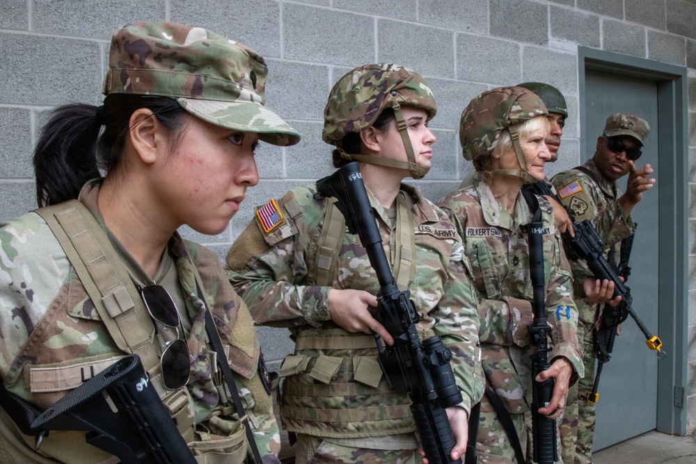 Five U.S. Army soldiers in Operational Camouflage Pattern (OCP) uniforms are positioned against a concrete block wall near a door. The group includes both male and female soldiers, several holding rifles, appearing to be in a training or operational briefing scenario. At least one soldier's name tape reading 'FOLKERTSMA' is partially visible.