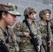Five U.S. Army soldiers in Operational Camouflage Pattern (OCP) uniforms are positioned against a concrete block wall near a door. The group includes both male and female soldiers, several holding rifles, appearing to be in a training or operational briefing scenario. At least one soldier's name tape reading 'FOLKERTSMA' is partially visible.