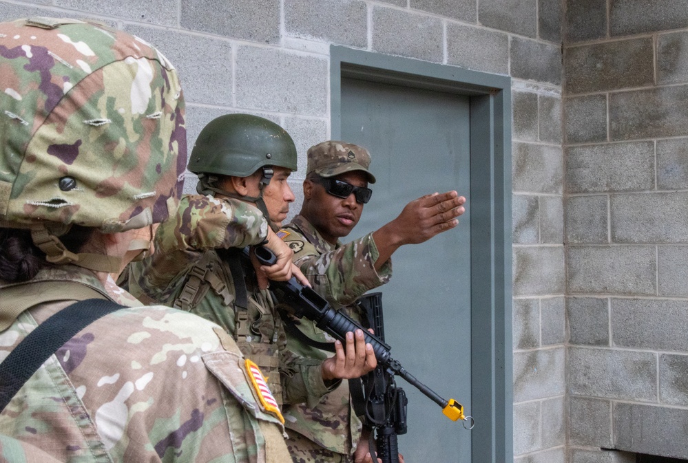 Three soldiers in US Army MultiCam (OCP) uniforms are conducting what appears to be a military training exercise involving building entry or room-clearing techniques. One soldier wearing sunglasses and a patrol cap appears to be an instructor gesturing toward a gray door in a cinder block structure. A second soldier holds a rifle with a yellow training device attached to the muzzle, indicating a simulated training environment.