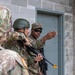 Three soldiers in US Army MultiCam (OCP) uniforms are conducting what appears to be a military training exercise involving building entry or room-clearing techniques. One soldier wearing sunglasses and a patrol cap appears to be an instructor gesturing toward a gray door in a cinder block structure. A second soldier holds a rifle with a yellow training device attached to the muzzle, indicating a simulated training environment.