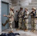 Four military personnel in OCP (Operational Camouflage Pattern) uniforms are conducting what appears to be a room-clearing or building-entry training exercise at a cinder block structure. One individual, appearing to be an instructor, is gesturing and providing guidance to three others who are holding training rifles. One trainee is positioned at the door, appearing to prepare for a breach entry.