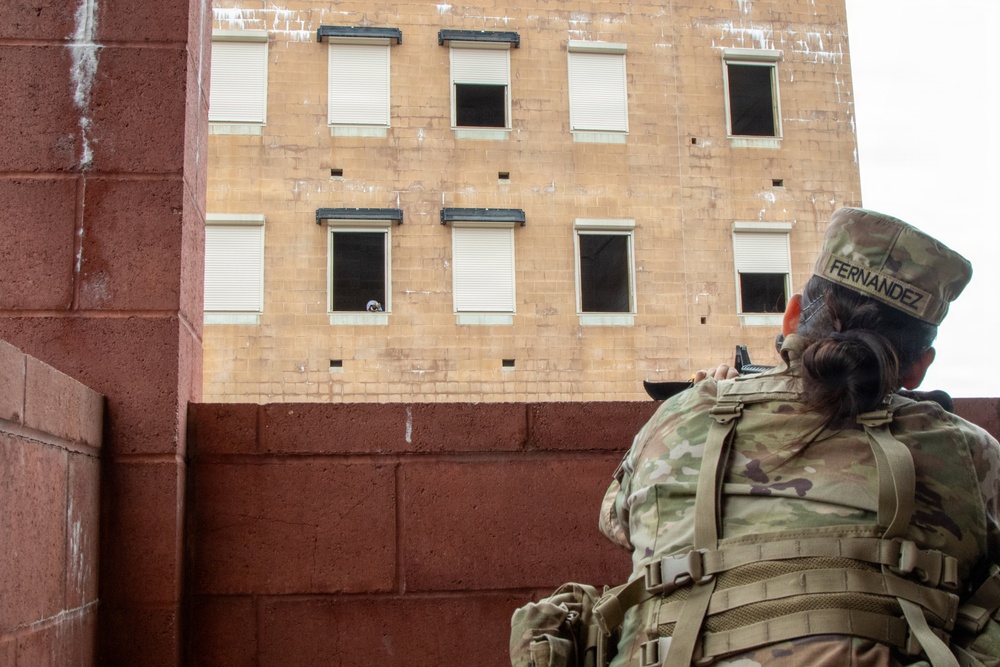 A soldier with the name tape 'FERNANDEZ' is seen from behind, wearing full multicam combat gear and a patrol cap, appearing to observe or provide security toward a multi-story training building. The building has multiple windows, some with roll-down shutters and some open, suggesting a military urban operations training facility. A figure is partially visible in one of the lower windows of the building.