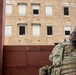 A soldier with the name tape 'FERNANDEZ' is seen from behind, wearing full multicam combat gear and a patrol cap, appearing to observe or provide security toward a multi-story training building. The building has multiple windows, some with roll-down shutters and some open, suggesting a military urban operations training facility. A figure is partially visible in one of the lower windows of the building.