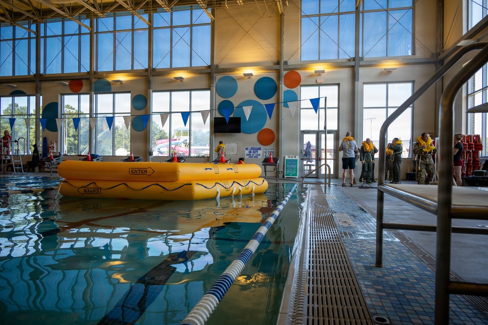 An indoor swimming pool facility is being used for what appears to be a military or emergency survival training exercise. A large yellow inflatable life raft labeled 'ENTER' is deployed in the pool, while several individuals wearing yellow life vests and military-style gear are gathered near the poolside. The facility features large windows, colorful circular wall decorations, and lane dividers typical of a recreational aquatic center.