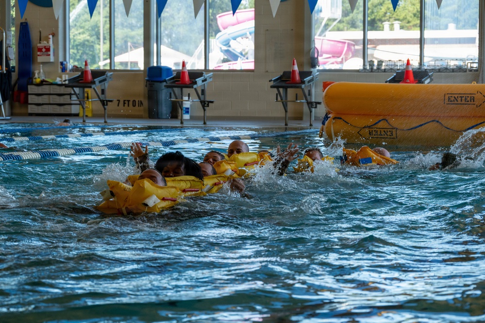 The image shows multiple individuals in an indoor swimming pool conducting what appears to be a water survival or rescue training exercise. Participants are wearing yellow life vests and are clustered together in the water near a large yellow inflatable life raft marked 'ENTER'. The setting is a recreational facility with lane markers, safety cones, and large windows overlooking an outdoor water park area.