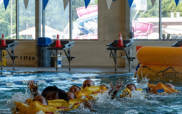 The image shows multiple individuals in an indoor swimming pool conducting what appears to be a water survival or rescue training exercise. Participants are wearing yellow life vests and are clustered together in the water near a large yellow inflatable life raft marked 'ENTER'. The setting is a recreational facility with lane markers, safety cones, and large windows overlooking an outdoor water park area.