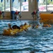 The image shows multiple individuals in an indoor swimming pool conducting what appears to be a water survival or rescue training exercise. Participants are wearing yellow life vests and are clustered together in the water near a large yellow inflatable life raft marked 'ENTER'. The setting is a recreational facility with lane markers, safety cones, and large windows overlooking an outdoor water park area.