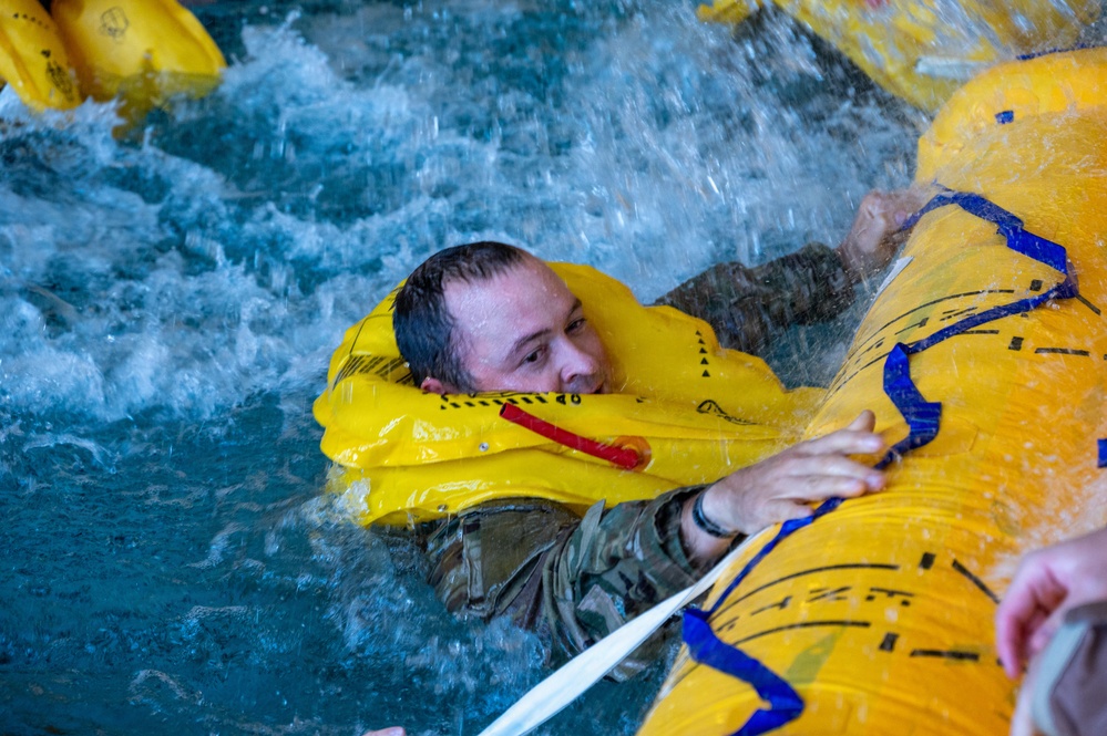A person wearing military camouflage clothing and an inflated yellow life preserver collar is in turbulent water, gripping a large yellow inflatable raft or life raft. The scene appears to be a water survival training exercise conducted in an indoor pool facility. Multiple yellow inflatable devices are visible in the churning water around the individual.