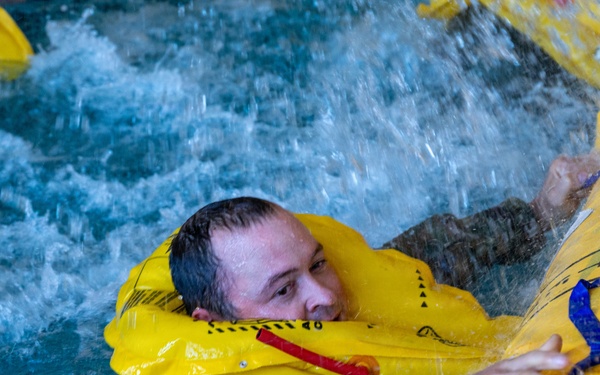 A person wearing military camouflage clothing and an inflated yellow life preserver collar is in turbulent water, gripping a large yellow inflatable raft or life raft. The scene appears to be a water survival training exercise conducted in an indoor pool facility. Multiple yellow inflatable devices are visible in the churning water around the individual.