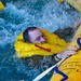 A person wearing military camouflage clothing and an inflated yellow life preserver collar is in turbulent water, gripping a large yellow inflatable raft or life raft. The scene appears to be a water survival training exercise conducted in an indoor pool facility. Multiple yellow inflatable devices are visible in the churning water around the individual.