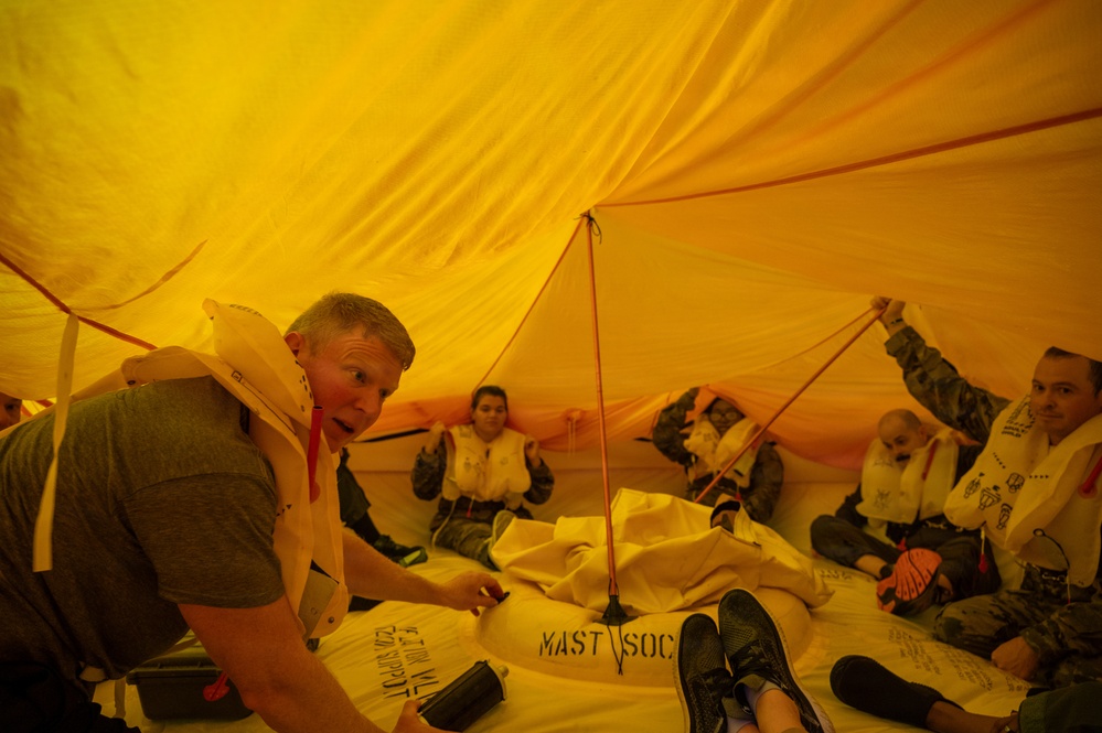 Several people are seated inside an inflated yellow life raft or survival craft, wearing life vests/personal flotation devices. The interior is bathed in orange-yellow light from the translucent canopy material. This appears to be a maritime or aviation survival training exercise.