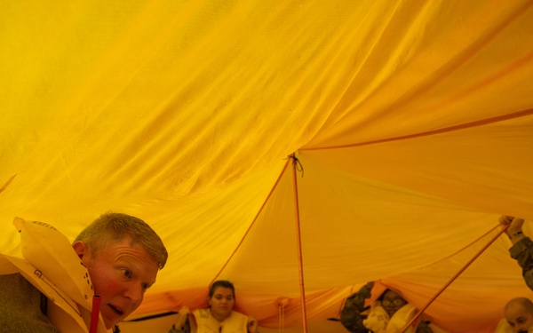 Several people are seated inside an inflated yellow life raft or survival craft, wearing life vests/personal flotation devices. The interior is bathed in orange-yellow light from the translucent canopy material. This appears to be a maritime or aviation survival training exercise.