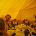 Several people are seated inside an inflated yellow life raft or survival craft, wearing life vests/personal flotation devices. The interior is bathed in orange-yellow light from the translucent canopy material. This appears to be a maritime or aviation survival training exercise.