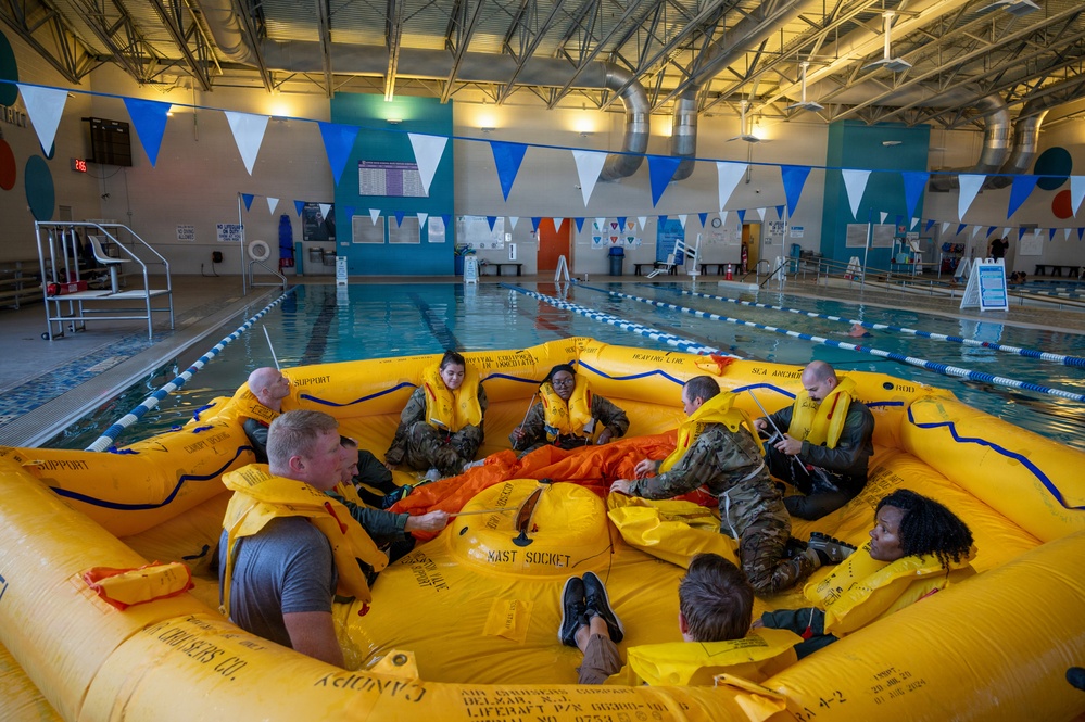 A group of military personnel in camouflage uniforms and yellow life vests are seated inside a large yellow inflatable life raft during a water survival training exercise conducted in an indoor swimming pool. The training appears to be part of a formal military survival course, with participants practicing raft procedures and equipment familiarization. The setting is a well-lit recreational indoor pool facility decorated with blue and white pennant banners.