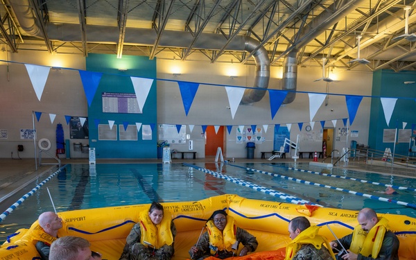 A group of military personnel in camouflage uniforms and yellow life vests are seated inside a large yellow inflatable life raft during a water survival training exercise conducted in an indoor swimming pool. The training appears to be part of a formal military survival course, with participants practicing raft procedures and equipment familiarization. The setting is a well-lit recreational indoor pool facility decorated with blue and white pennant banners.