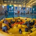 A group of military personnel in camouflage uniforms and yellow life vests are seated inside a large yellow inflatable life raft during a water survival training exercise conducted in an indoor swimming pool. The training appears to be part of a formal military survival course, with participants practicing raft procedures and equipment familiarization. The setting is a well-lit recreational indoor pool facility decorated with blue and white pennant banners.