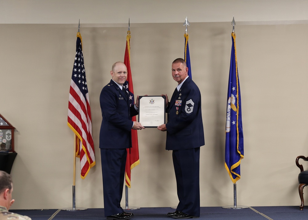 Two U.S. Air Force personnel are standing on a stage during a formal military ceremony, jointly holding a framed certificate or commendation document. The individual on the right appears to be a senior non-commissioned officer, likely a Chief Master Sergeant, receiving recognition. Four flags are displayed in the background, including the U.S. flag, a state flag, and Air Force-related flags.