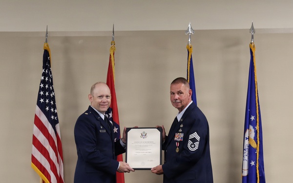 Two U.S. Air Force personnel are standing on a stage during a formal military ceremony, jointly holding a framed certificate or commendation document. The individual on the right appears to be a senior non-commissioned officer, likely a Chief Master Sergeant, receiving recognition. Four flags are displayed in the background, including the U.S. flag, a state flag, and Air Force-related flags.
