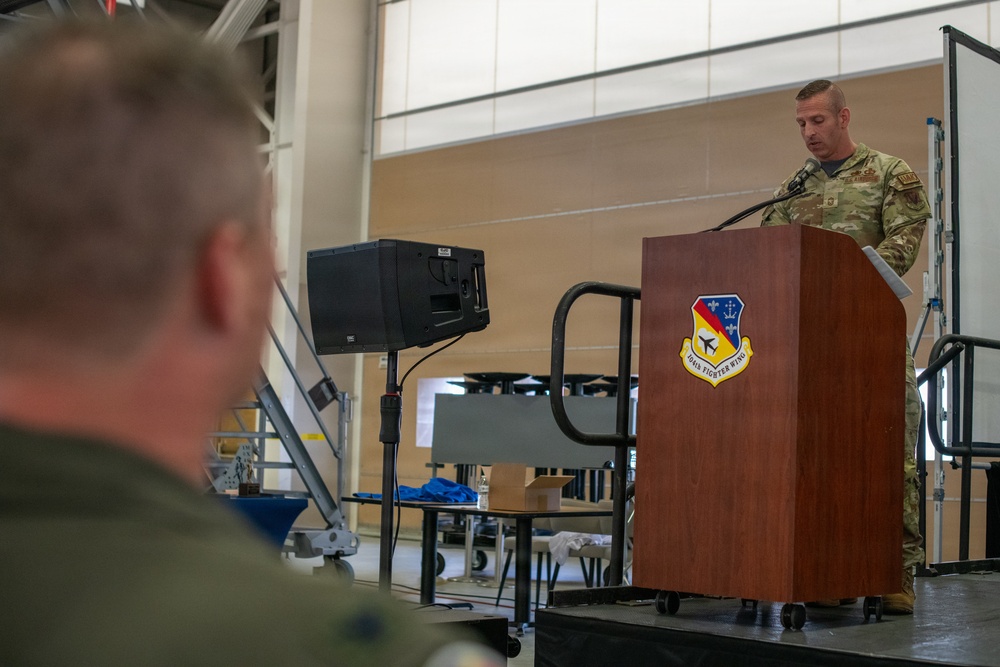 A military personnel in OCP (Operational Camouflage Pattern) uniform is speaking at a wooden podium bearing the insignia of the 134th Fighter Wing during what appears to be an official ceremony or briefing. The event is taking place inside a large hangar facility with another service member visible in the foreground observing the speaker. A portable PA speaker on a stand is positioned near the stage.