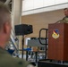 A military personnel in OCP (Operational Camouflage Pattern) uniform is speaking at a wooden podium bearing the insignia of the 134th Fighter Wing during what appears to be an official ceremony or briefing. The event is taking place inside a large hangar facility with another service member visible in the foreground observing the speaker. A portable PA speaker on a stand is positioned near the stage.