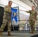 Two military personnel in OCP (Operational Camouflage Pattern) uniforms are on a stage during what appears to be a formal military ceremony. One soldier is unfurling or casing a blue guidon flag bearing the insignia of the 104th Maintenance Group (104 MXG) and 104th Munitions Squadron (104 MUNS). The event appears to be a unit activation, inactivation, or change of command ceremony held inside a military hangar.