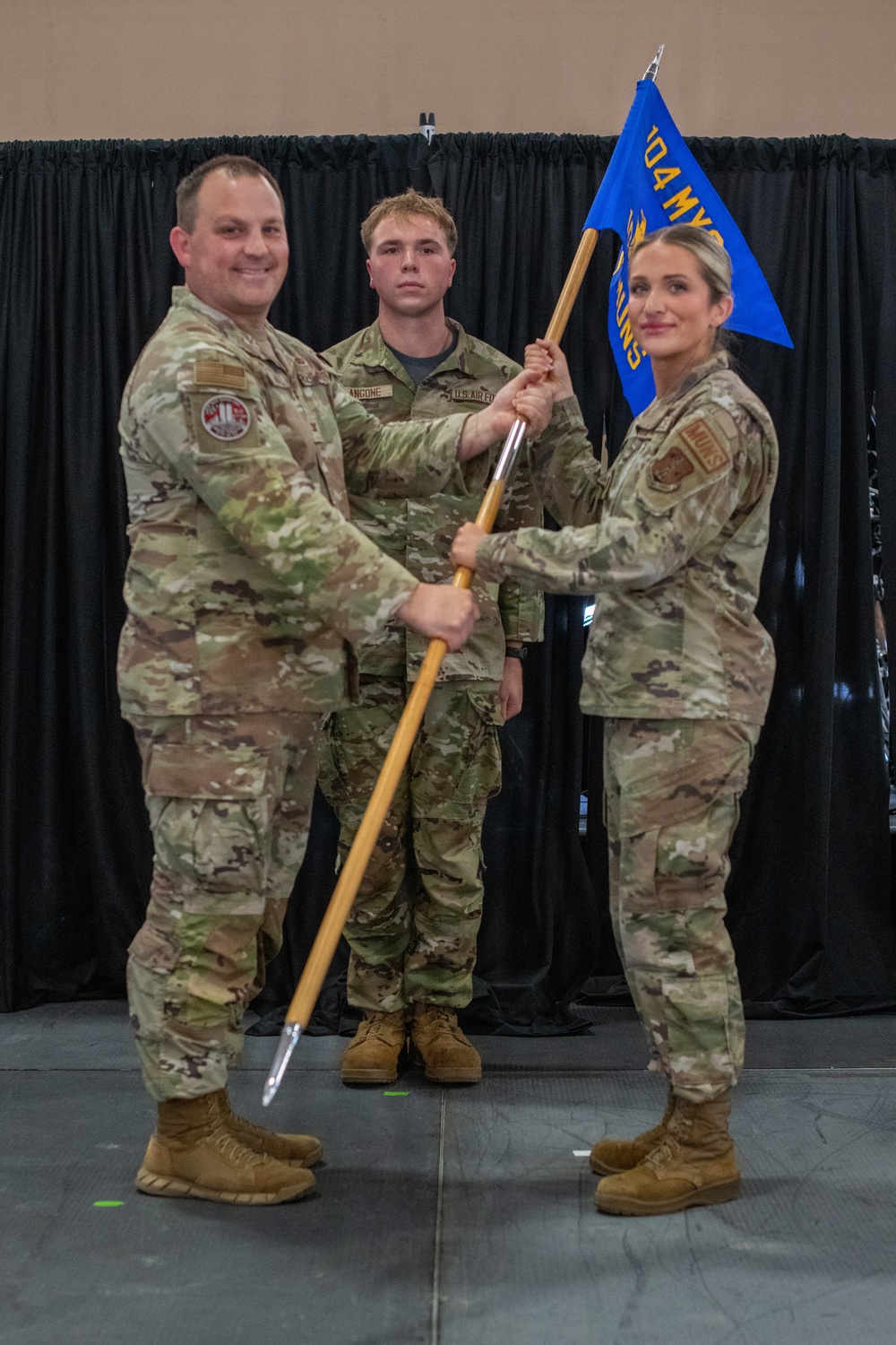 Three U.S. Air Force personnel in OCP (Operational Camouflage Pattern) uniforms are participating in a guidon passing ceremony, likely a change of command or activation event. Two individuals are jointly holding a blue guidon staff while a third stands at attention in the background. The blue flag bears the text '104 MXS' (104th Maintenance Squadron) in gold lettering.