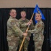 Three U.S. Air Force personnel in OCP (Operational Camouflage Pattern) uniforms are participating in a guidon passing ceremony, likely a change of command or activation event. Two individuals are jointly holding a blue guidon staff while a third stands at attention in the background. The blue flag bears the text '104 MXS' (104th Maintenance Squadron) in gold lettering.