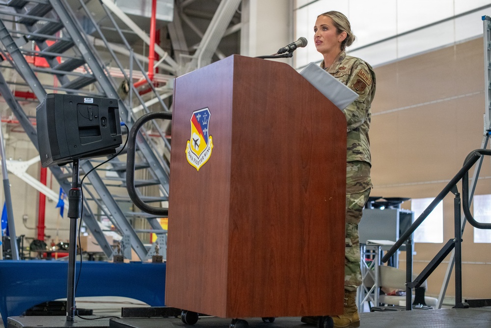 A female U.S. Air Force service member in OCP (Operational Camouflage Pattern) uniform is speaking at a wooden podium bearing a military unit insignia, identified as the 'Fighter Wing' emblem. The setting appears to be inside a military hangar or large maintenance facility, with industrial scaffolding and equipment visible in the background. A teleprompter or monitor on a stand is positioned to her left.