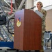 A female U.S. Air Force service member in OCP (Operational Camouflage Pattern) uniform is speaking at a wooden podium bearing a military unit insignia, identified as the 'Fighter Wing' emblem. The setting appears to be inside a military hangar or large maintenance facility, with industrial scaffolding and equipment visible in the background. A teleprompter or monitor on a stand is positioned to her left.