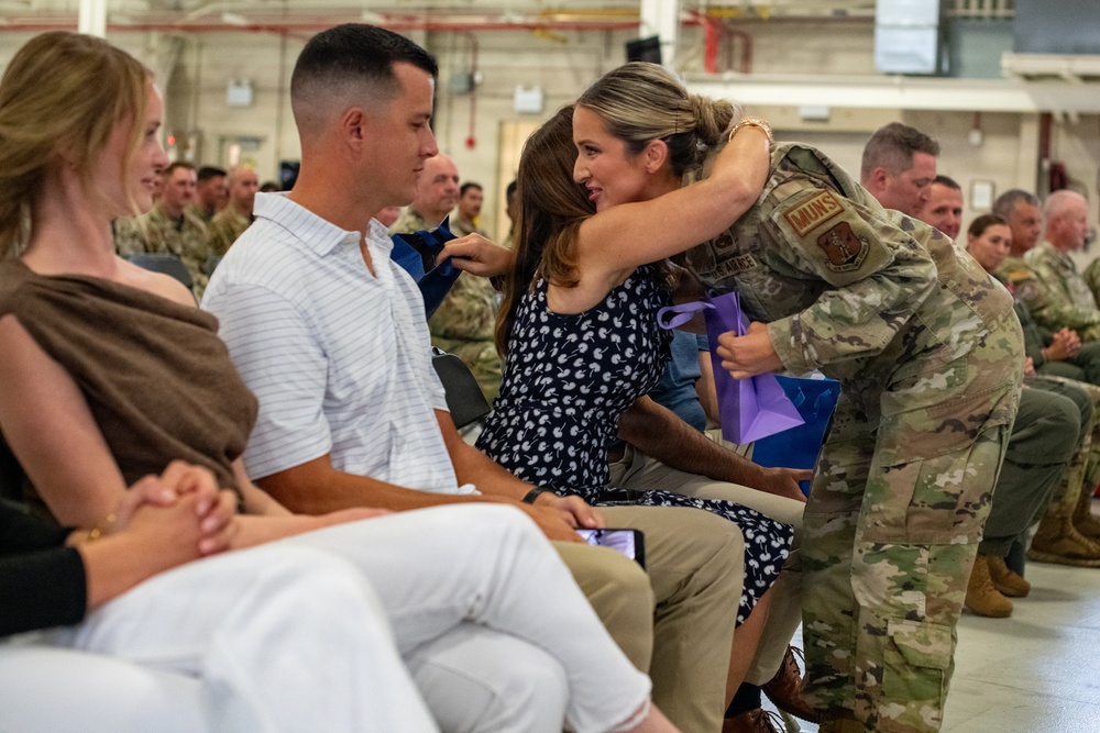 A military ceremony taking place in what appears to be a hangar or large indoor facility. A service member in OCP (Operational Camouflage Pattern) uniform is leaning down to embrace a seated civilian woman, while holding a purple gift bag. Rows of uniformed military personnel are visible in the background, suggesting a formal recognition or farewell ceremony.