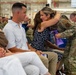 A military ceremony taking place in what appears to be a hangar or large indoor facility. A service member in OCP (Operational Camouflage Pattern) uniform is leaning down to embrace a seated civilian woman, while holding a purple gift bag. Rows of uniformed military personnel are visible in the background, suggesting a formal recognition or farewell ceremony.