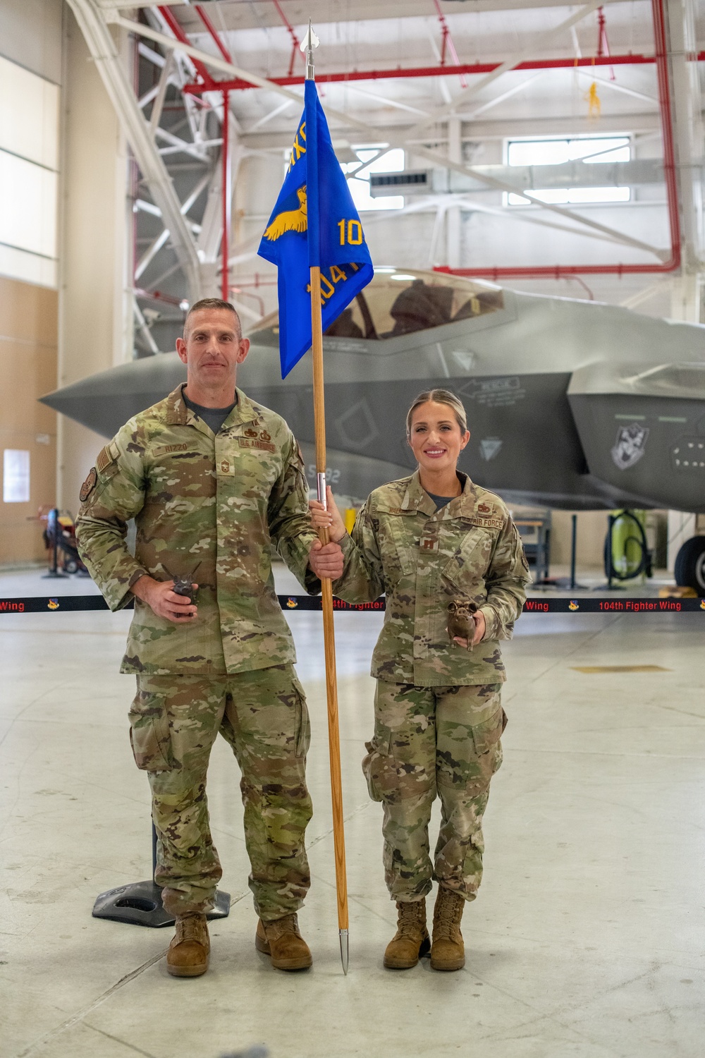 Two U.S. Air Force personnel in OCP (Operational Camouflage Pattern) uniforms stand in an aircraft hangar in front of an F-35 fighter jet. The male service member on the left holds a blue guidon flag bearing the number '104' and an eagle emblem, consistent with the 104th Fighter Wing. A barrier tape reading '104th Fighter Wing' is visible in the foreground.