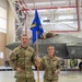 Two U.S. Air Force personnel in OCP (Operational Camouflage Pattern) uniforms stand in an aircraft hangar in front of an F-35 fighter jet. The male service member on the left holds a blue guidon flag bearing the number '104' and an eagle emblem, consistent with the 104th Fighter Wing. A barrier tape reading '104th Fighter Wing' is visible in the foreground.