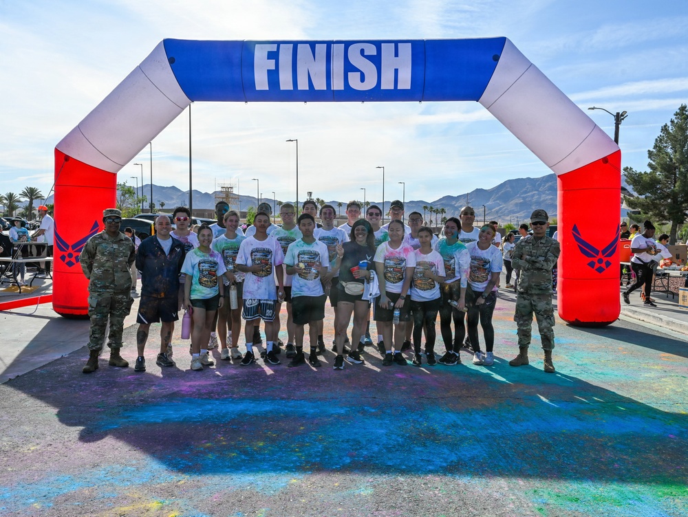A group of approximately 20 participants pose under an inflatable 'FINISH' arch at what appears to be a color run or 5K event on a military installation, indicated by the U.S. Air Force logos on the arch. Two individuals in military camouflage uniforms (ACUs) flank the group, while the majority wear matching white event t-shirts stained with colorful powder. The ground beneath them is visibly coated with blue, green, and pink color powder residue.
