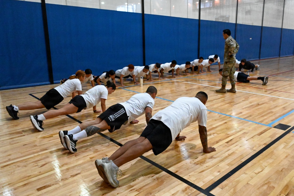 A large group of individuals in white t-shirts and black shorts are performing push-ups in a gymnasium with a hardwood basketball court floor. A single person in military camouflage uniform (OCP pattern) stands and observes the group, suggesting a military physical training or recruitment assessment session. The participants are arranged in a diagonal line formation across the court.