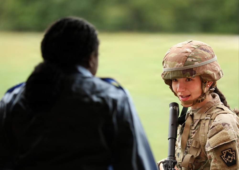 A U.S. Army soldier in full Operational Camouflage Pattern (OCP) uniform and combat helmet is engaged in conversation with a civilian or non-uniformed individual whose back is to the camera. The soldier is holding what appears to be a rifle and has a braided hairstyle visible beneath the helmet. The setting appears to be an outdoor field or training area with green grass and trees in the background.