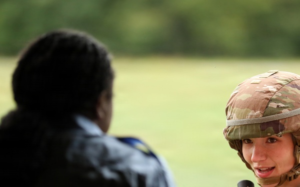 A U.S. Army soldier in full Operational Camouflage Pattern (OCP) uniform and combat helmet is engaged in conversation with a civilian or non-uniformed individual whose back is to the camera. The soldier is holding what appears to be a rifle and has a braided hairstyle visible beneath the helmet. The setting appears to be an outdoor field or training area with green grass and trees in the background.