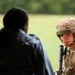 A U.S. Army soldier in full Operational Camouflage Pattern (OCP) uniform and combat helmet is engaged in conversation with a civilian or non-uniformed individual whose back is to the camera. The soldier is holding what appears to be a rifle and has a braided hairstyle visible beneath the helmet. The setting appears to be an outdoor field or training area with green grass and trees in the background.